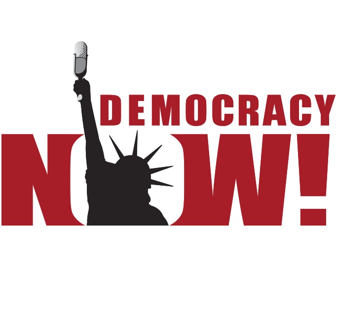 The image features the logo for “Democracy Now!” The design includes the silhouette of the Statue of Liberty holding a microphone in place of the torch. The words “DEMOCRACY NOW!” are displayed prominently in bold red letters, with “DEMOCRACY” above and “NOW!” below the statue’s arm. This logo represents the independent news program known for its progressive and critical reporting on political and social issues.