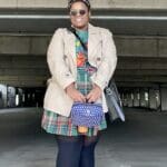 In the photo, Jalisa Bass stands confidently, smiling while wearing a stylish ensemble. Her outfit features a plaid skirt, a graphic t-shirt, and a beige trench coat. She accessorizes with round sunglasses, a vibrant scarf tied around her hair, a blue patterned handbag, and bright pink high-top sneakers. The setting appears to be an urban parking garage, providing a casual yet unique backdrop to her bold and eclectic style.