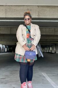 In the photo, Jalisa Bass stands confidently, smiling while wearing a stylish ensemble. Her outfit features a plaid skirt, a graphic t-shirt, and a beige trench coat. She accessorizes with round sunglasses, a vibrant scarf tied around her hair, a blue patterned handbag, and bright pink high-top sneakers. The setting appears to be an urban parking garage, providing a casual yet unique backdrop to her bold and eclectic style.