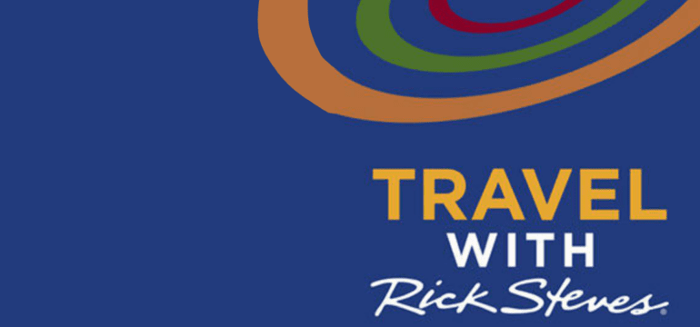 The image is a logo for the program “Travel with Rick Steves.” The background is a deep blue color. At the top, there are three curved lines in red, green, and brown, resembling stylized travel paths or trails, forming an abstract, circular pattern. Below these lines, the text “TRAVEL WITH Rick Steves” is prominently displayed. The word “TRAVEL” is in bold, uppercase yellow letters. The word “WITH” is in smaller, bold, uppercase white letters underneath “TRAVEL.” Below “WITH,” the name “Rick Steves” is written in a white, cursive font that resembles a signature. The overall design is clean and modern, with the curved lines adding a dynamic, travel-oriented feel to the logo. The text is clear and easy to read, with the deep blue background providing strong contrast.