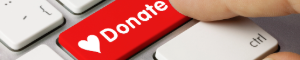 The image shows a red keyboard key labeled “Donate” with a heart symbol next to it. A finger is poised to press the key. This image serves as a button; clicking it will take you to the KBCS Donate page at https://staging-kbcsfm.kinsta.cloud/donate/.