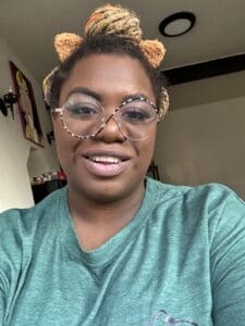 91.3 KBCS Jalisa Bass The image shows Jalisa smiling warmly at the camera, wearing a pair of stylish, large, round glasses. Her hair is styled in a bun with a headband that has orange and brown accents. She is wearing a green shirt, and the background shows part of a cozy room with artwork visible on the wall. The soft lighting highlights her friendly and approachable demeanor.