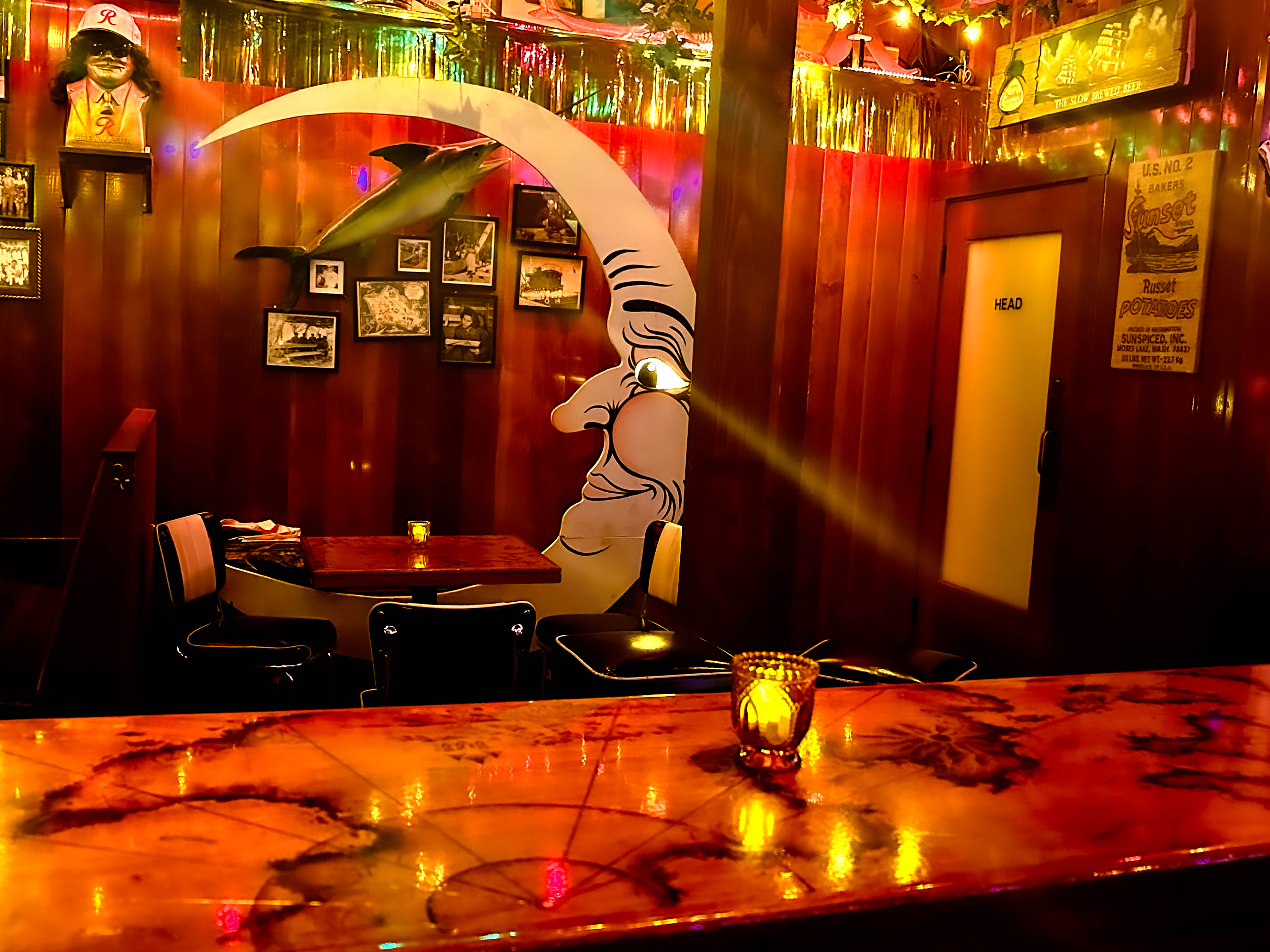 The image shows a cozy, dimly lit area of a tavern, likely a booth seating section with a whimsical mural of a crescent moon with a face looking toward a small table. The wooden walls are adorned with framed photos and decorations, including a mounted fish. The lighting is warm, casting an inviting glow from small candles on the tables and bar. A door marked “HEAD” can be seen in the background. The overall ambiance reflects a nostalgic, quirky vibe with a blend of retro decor, making it an intimate space for gatherings.