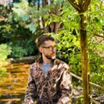 Realistic photograph of a white disabled non-binary person sitting by a pond under a pagoda in a garden. This person is wearing glasses, has short dark brown hair, and is wearing a fuzzy light brown paisley print sweater