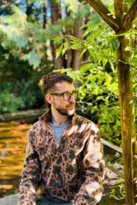 Realistic photograph of a white disabled non-binary person sitting by a pond under a pagoda in a garden. This person is wearing glasses, has short dark brown hair, and is wearing a fuzzy light brown paisley print sweater