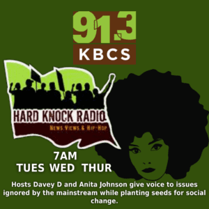 The image is a promotional graphic for the radio show Hard Knock Radio on 91.3 KBCS. It has a green background with the Hard Knock Radio logo prominently featured. The logo includes a silhouette of protestors holding signs, set against a green and black background. Below, there is an Afro-style silhouette of a woman’s face. The text on the image reads, “91.3 KBCS Hard Knock Radio, 7 AM, Tuesday, Wednesday, Thursday. Hosts Davey D and Anita Johnson give voice to issues ignored by the mainstream while planting seeds for social change.” The overall tone reflects activism and social justice themes.