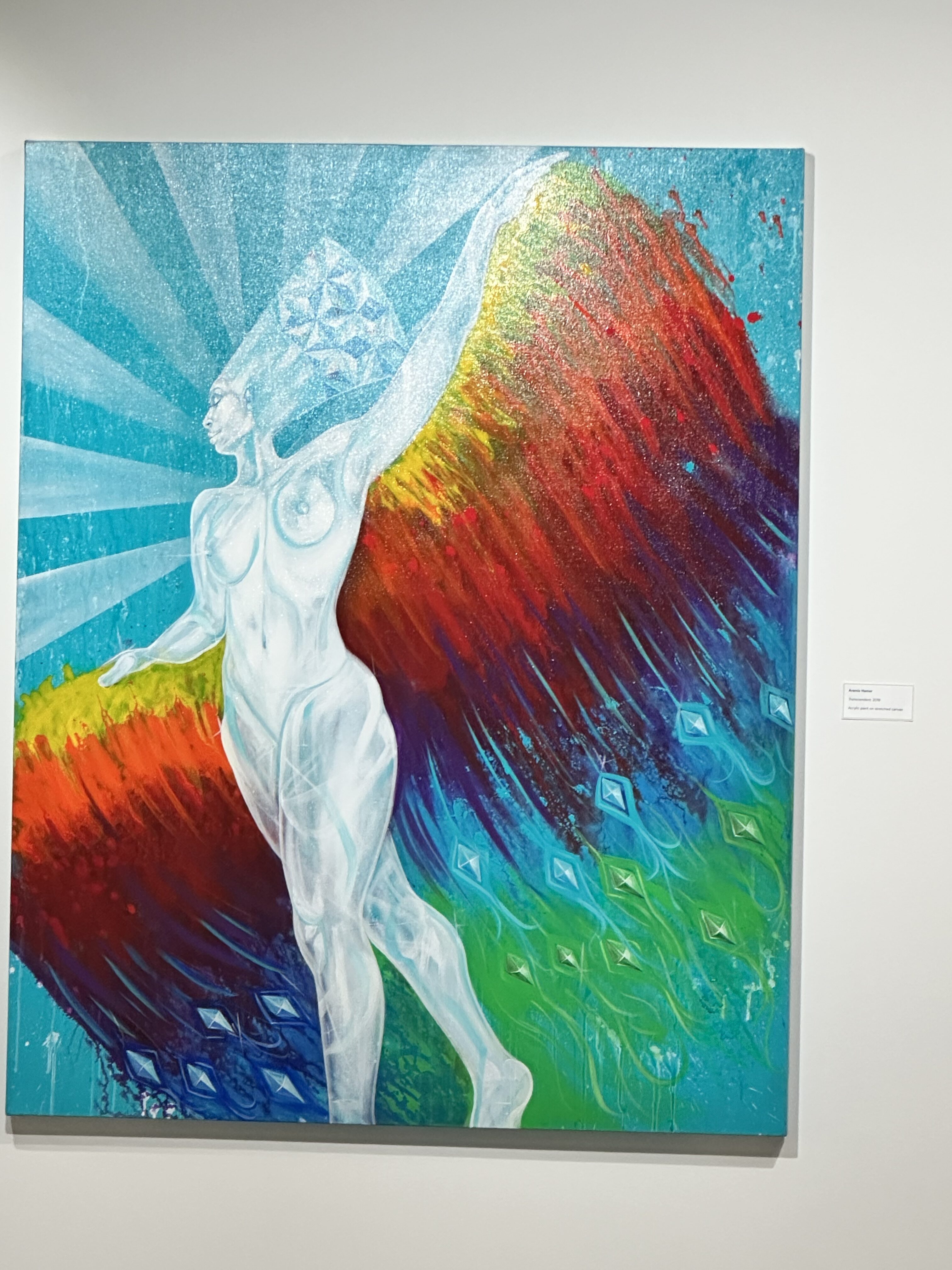 The attached image features a colorful and vibrant painting that depicts a powerful, ethereal figure. The figure, set against a radiant background of blue, green, red, and orange hues, appears to be floating or in mid-motion, with one arm outstretched. The figure’s body is rendered in a semi-transparent, glowing white with geometric patterns on the head, suggesting a sense of otherworldliness or transcendence. The vibrant colors and dynamic brushstrokes evoke a feeling of energy and freedom, while the composition blends elements of the surreal with abstract expression. The painting radiates a sense of movement, empowerment, and the blending of nature with the human form.